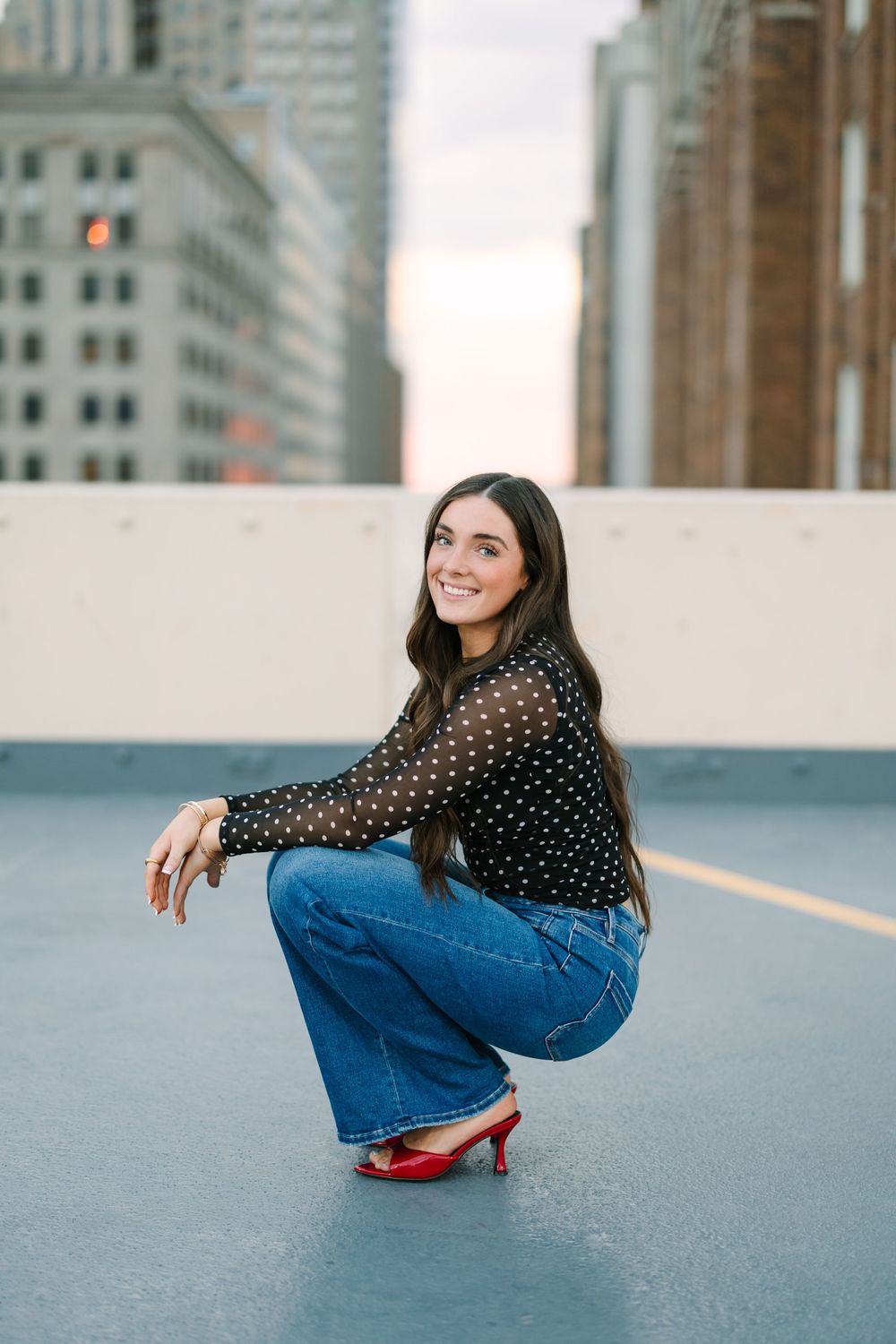 Young woman in polka dot top and jeans crouching on rooftop with city skyline at sunset.