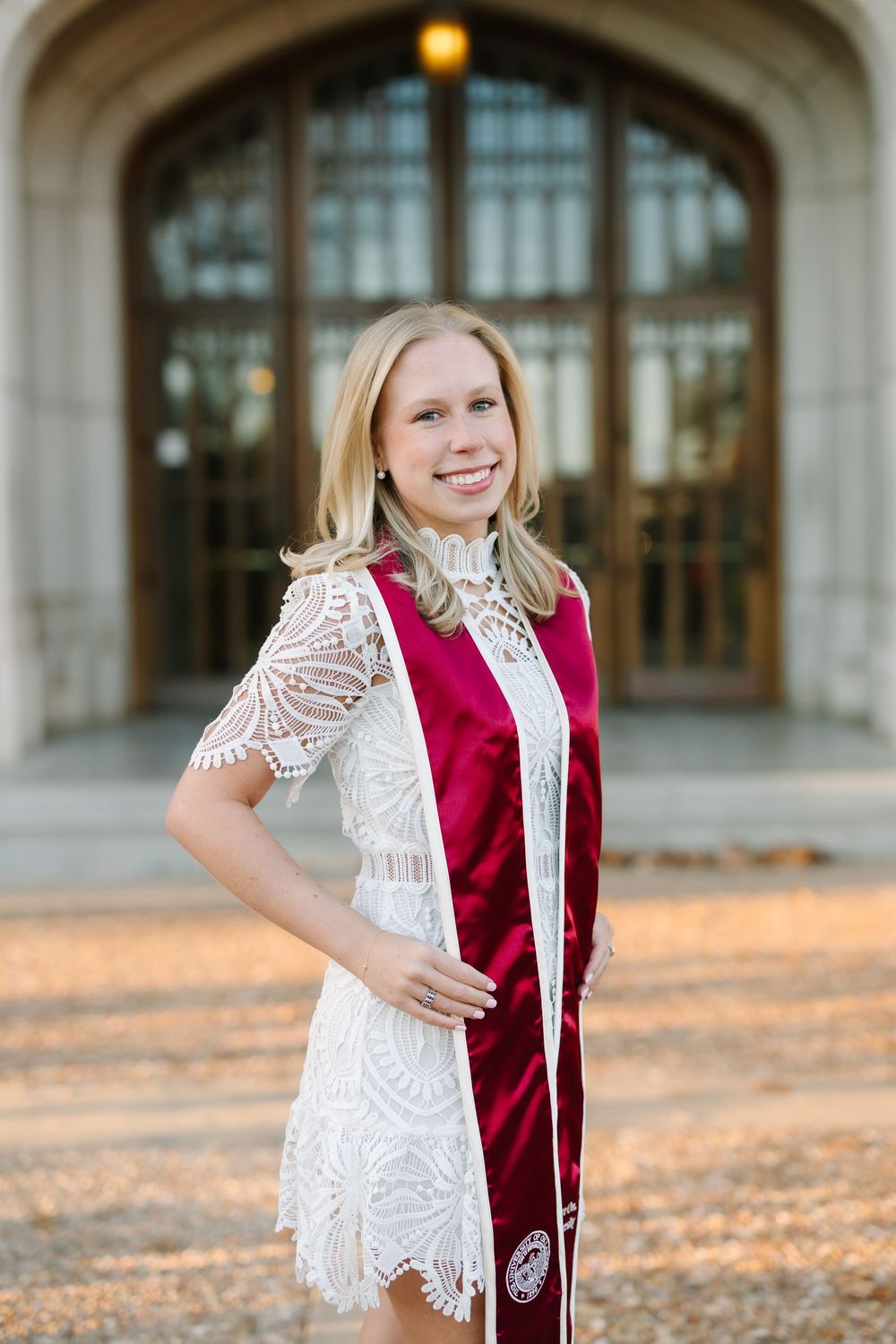 Smiling blonde woman in white lace dress and maroon graduation stole posing outdoors near an arched stone building.