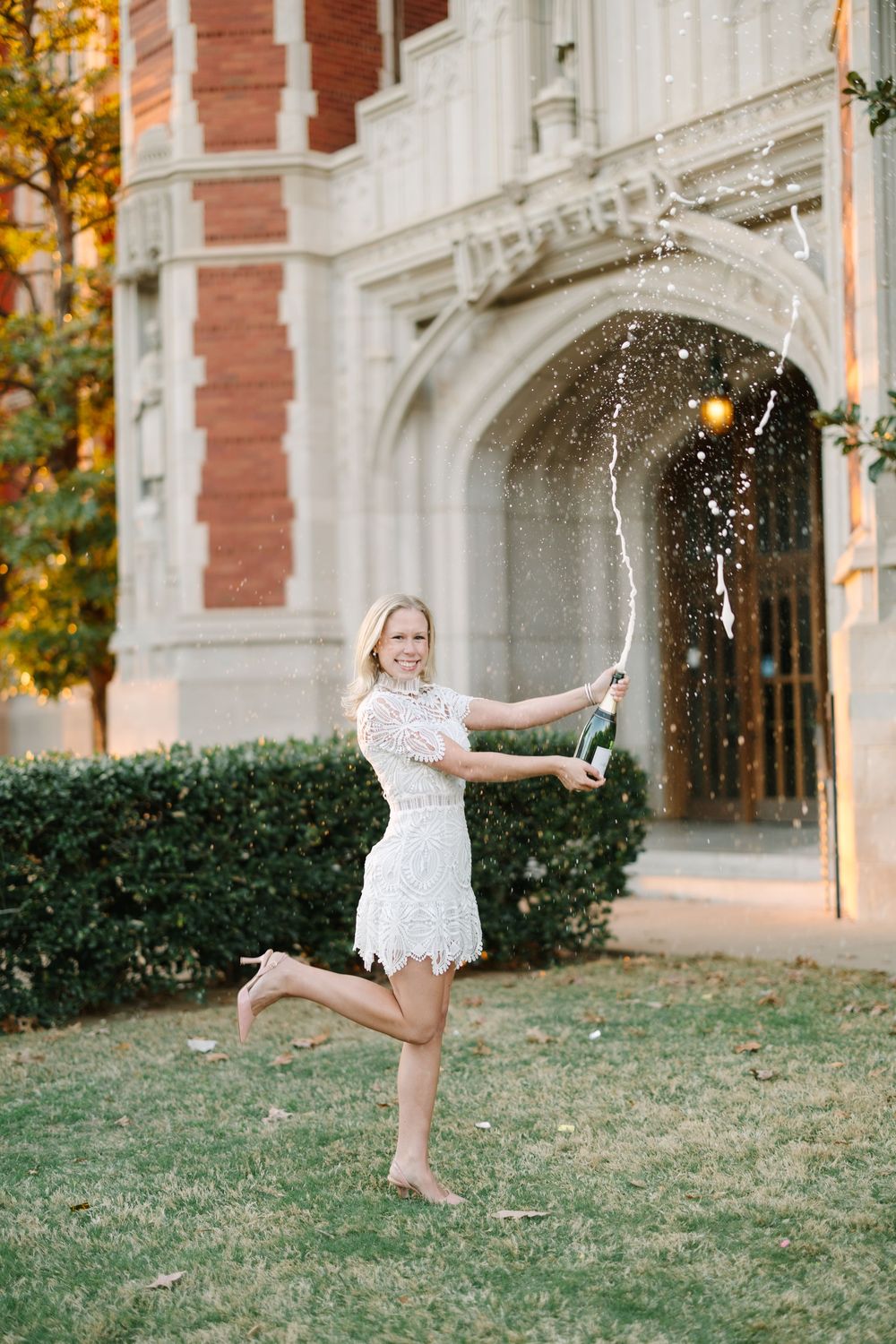 Woman in white lace dress celebrates outdoors, popping champagne bottle near brick building on green lawn.