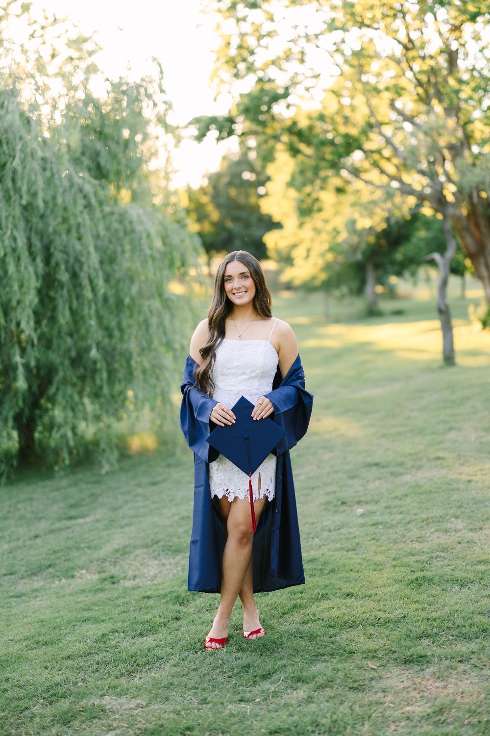Young woman in white dress and navy graduation gown holds cap outdoors on green lawn with trees.