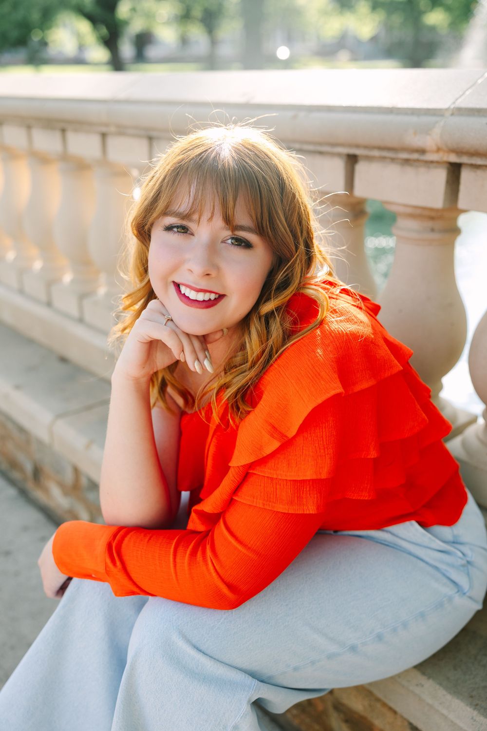 Person in bright orange ruffled top smiles while resting on gray fabric in sunlit outdoor setting.