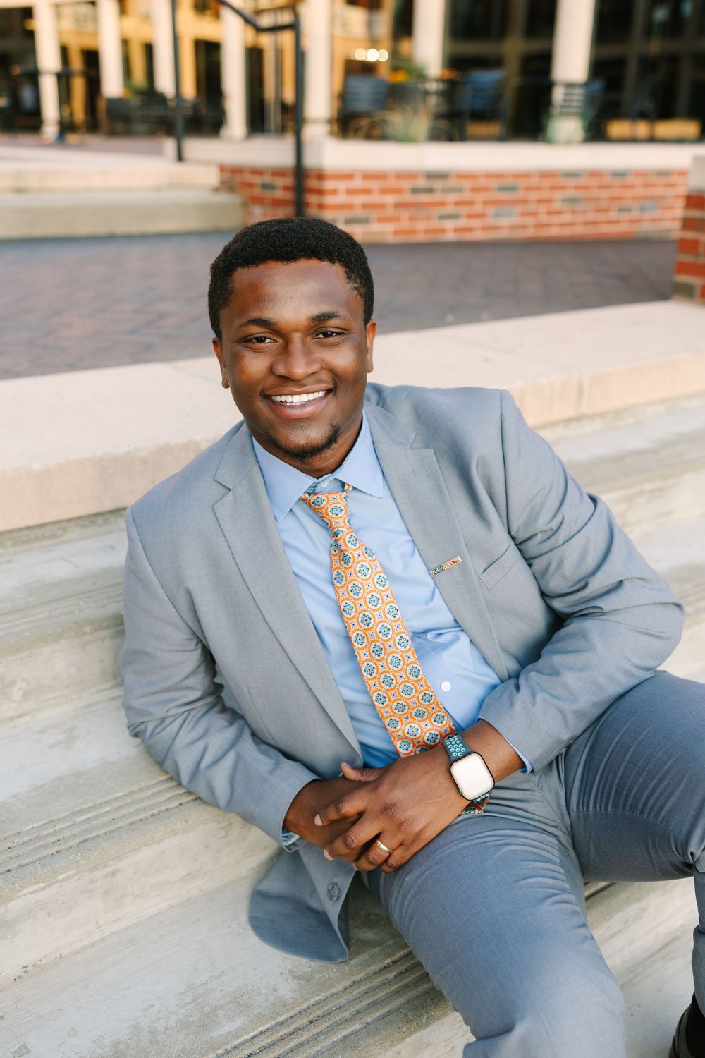 Professional wearing a light gray suit and yellow patterned tie sits casually on outdoor steps of brick building.