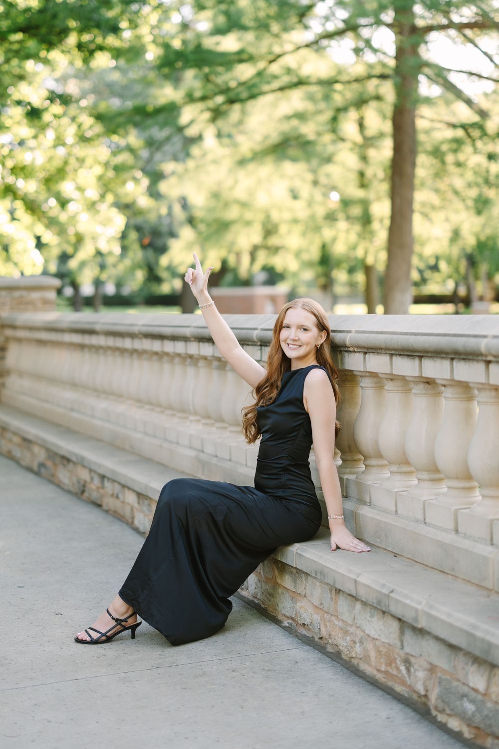 Young woman in black jumpsuit sitting on stone steps outdoors, smiling and raising her hand in a park setting.