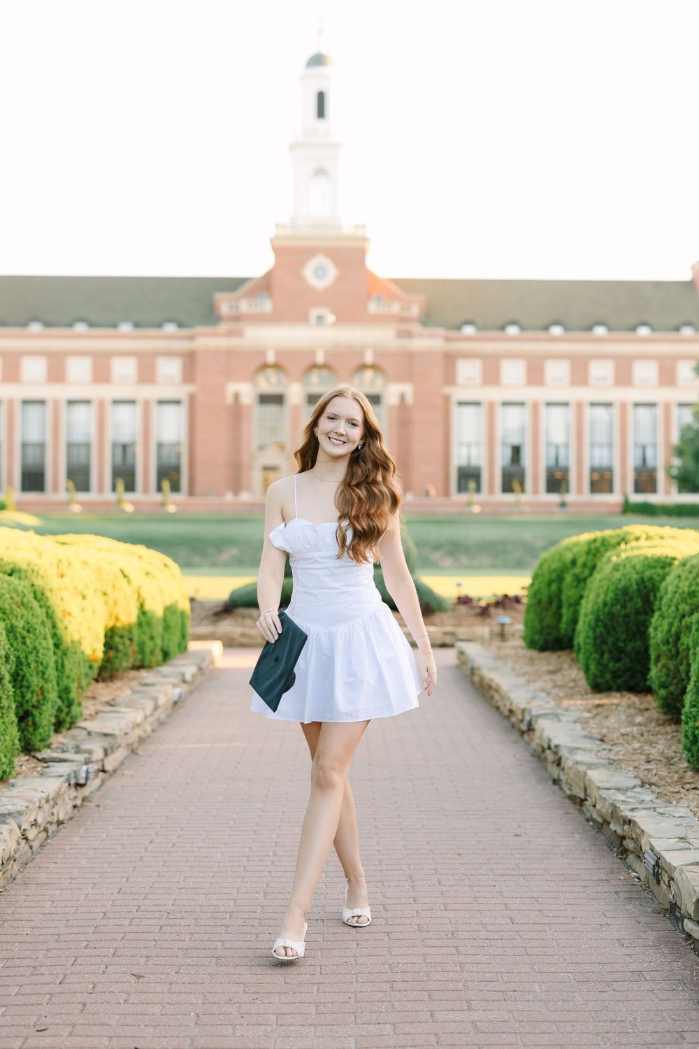 Young woman in white off-shoulder dress walks along brick path in front of red brick university building.
