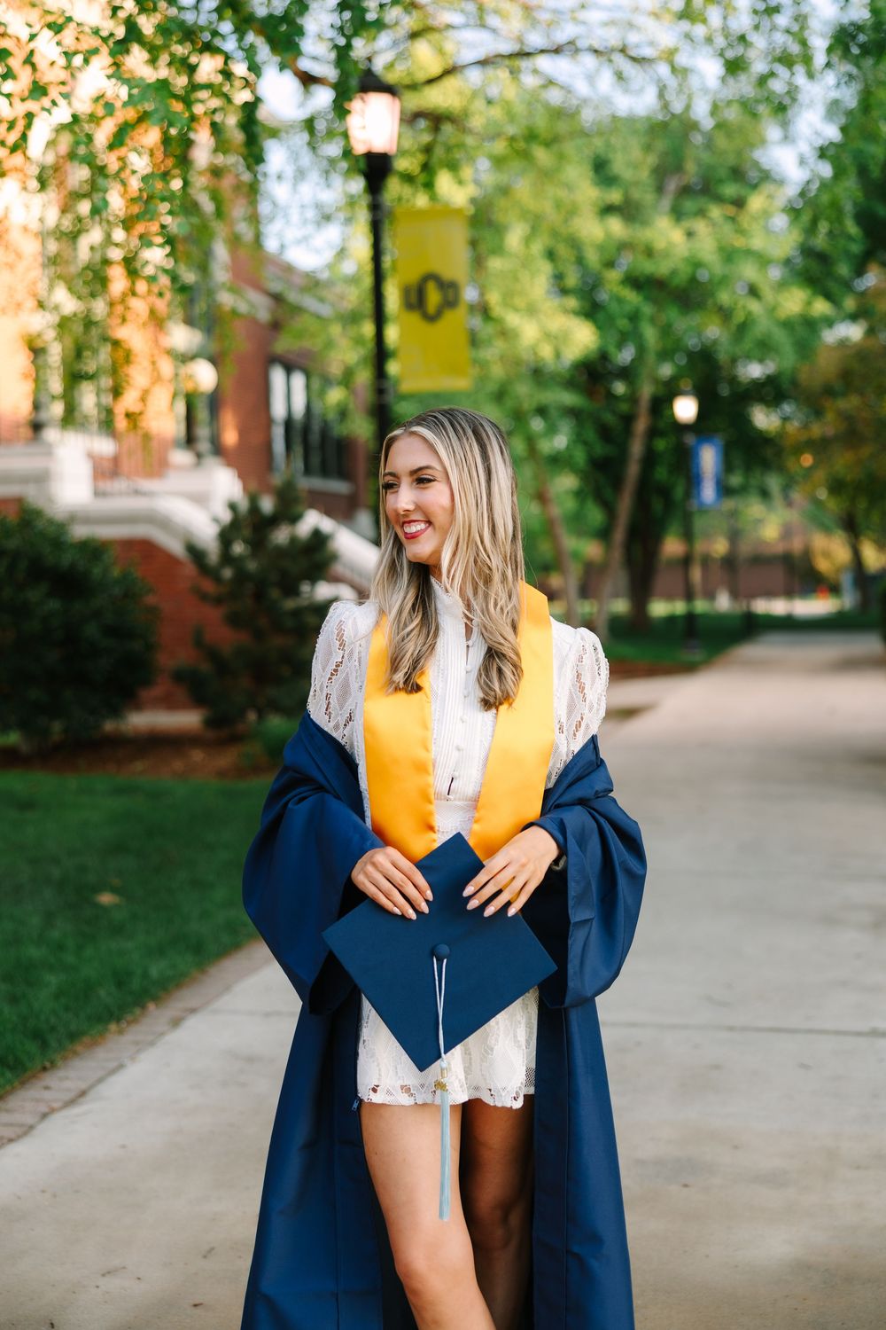Smiling female graduate in navy cap and gown with gold stole poses on a tree-lined campus walkway.
