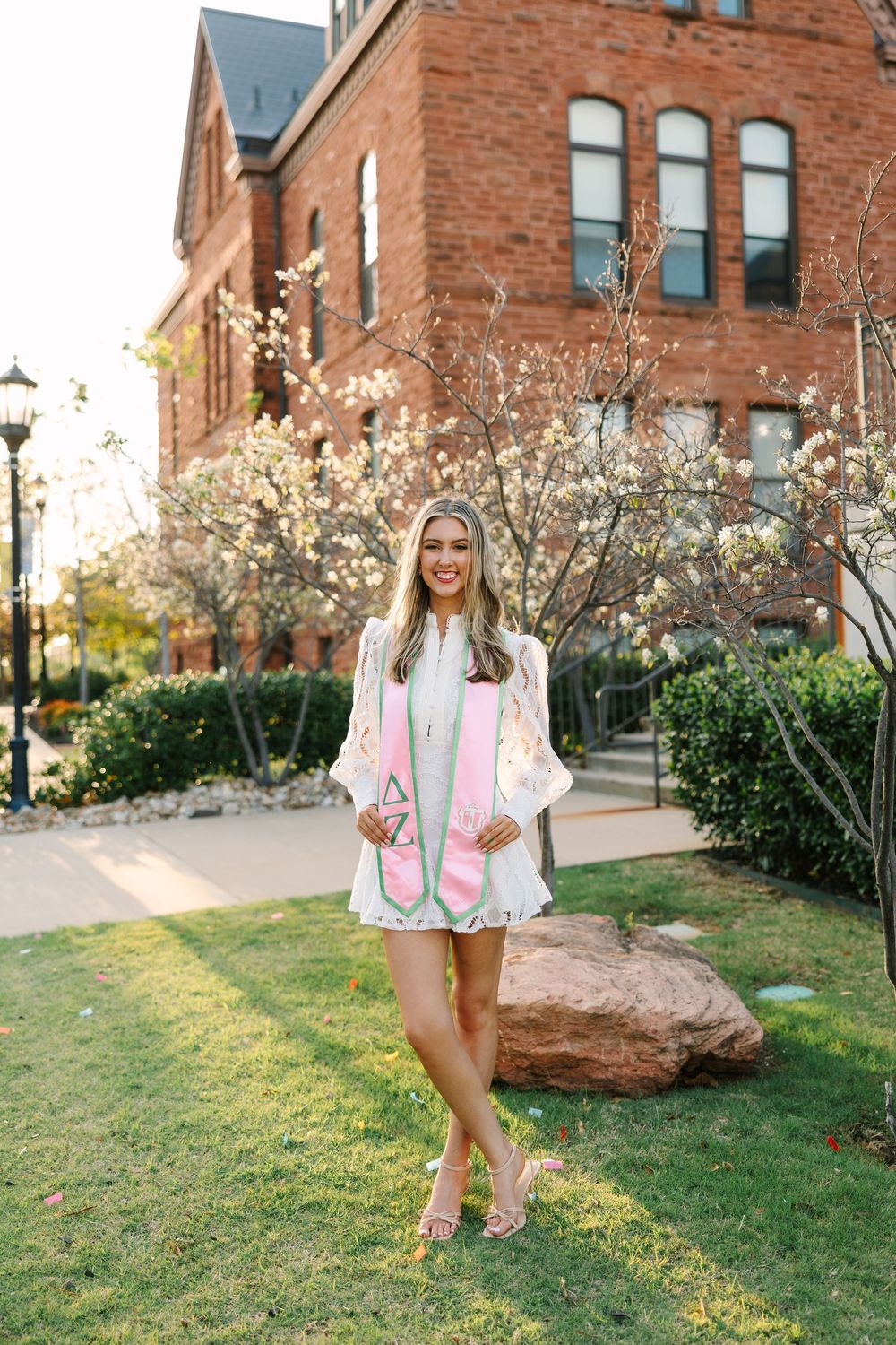 Smiling graduate in white gown and pink stole poses outdoors near brick building with blooming trees.