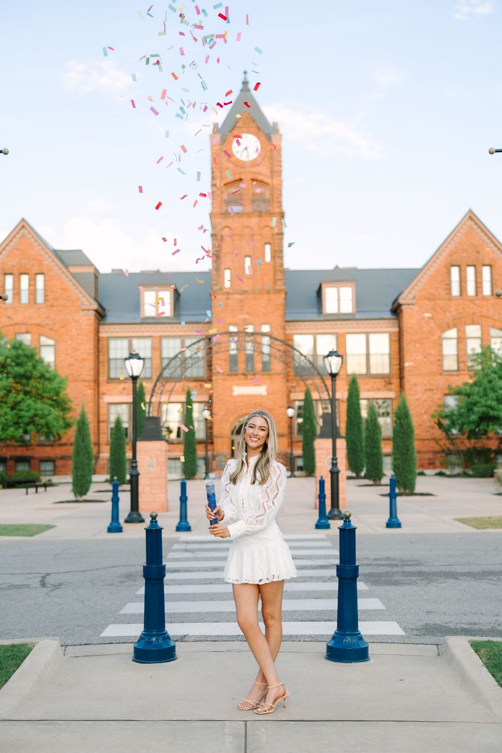 Young woman in white dress celebrates with confetti cannon in front of a red brick clock tower building on a sunny day.