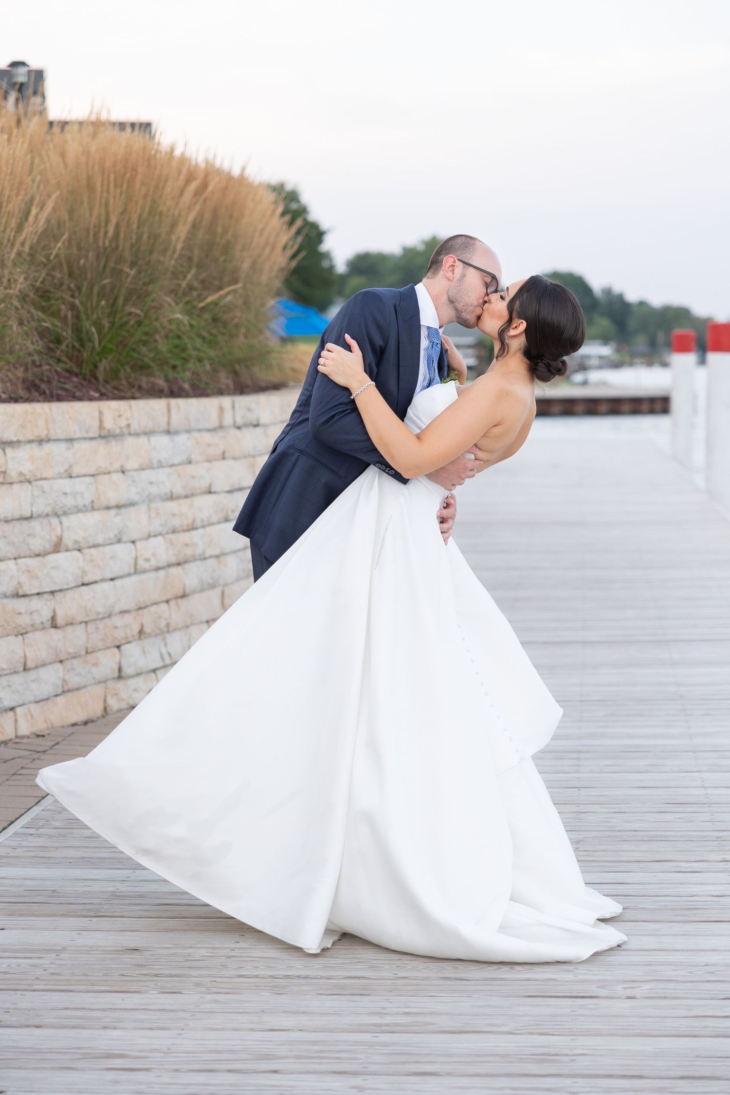 Newlywed couple shares romantic kiss on wooden boardwalk while bride's white dress flows in the breeze by the water. By Rachel Martinez.
