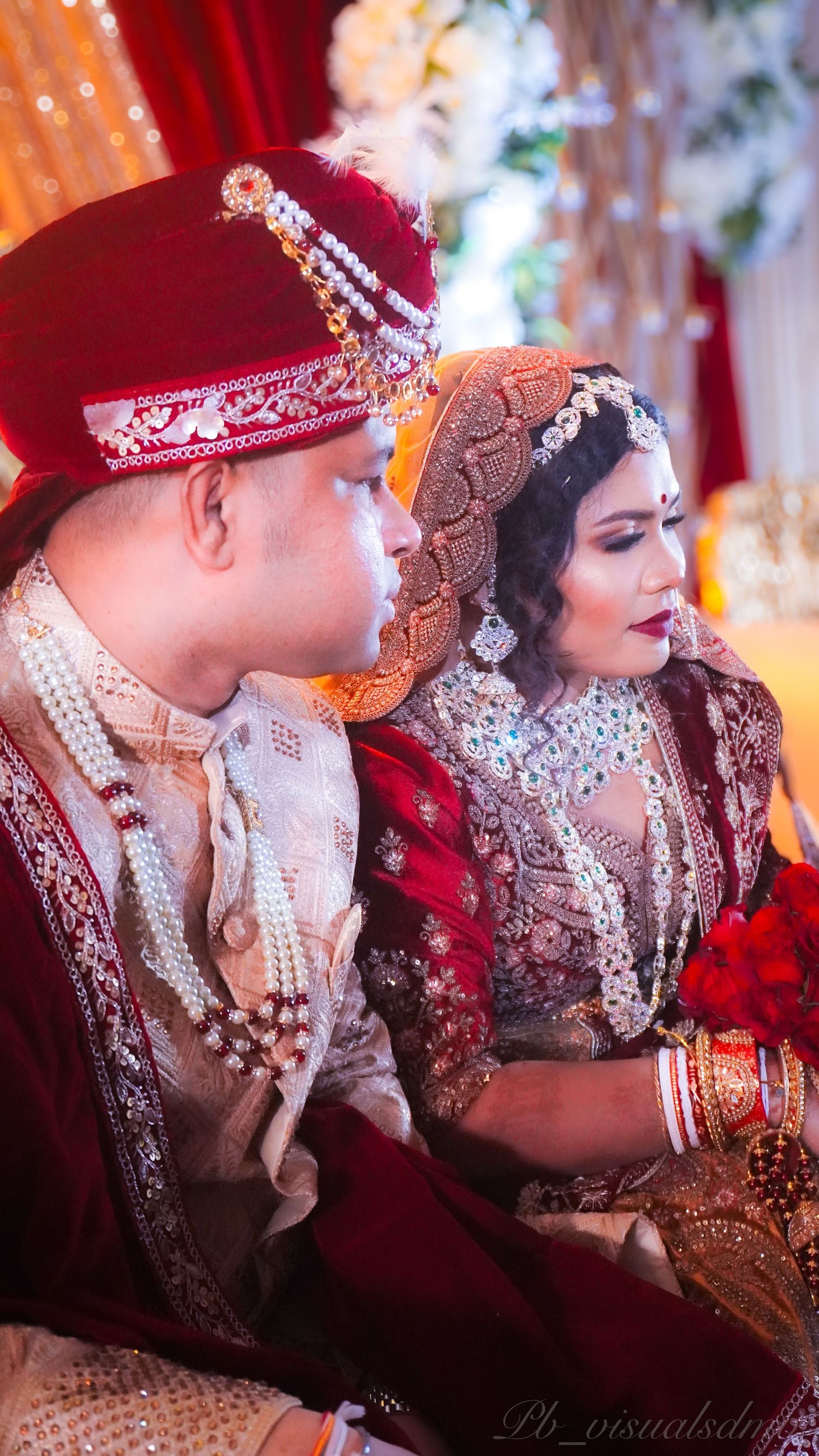 South Asian bride and groom in traditional red and gold wedding attire, captured during ceremony with intricate jewelry and décor.