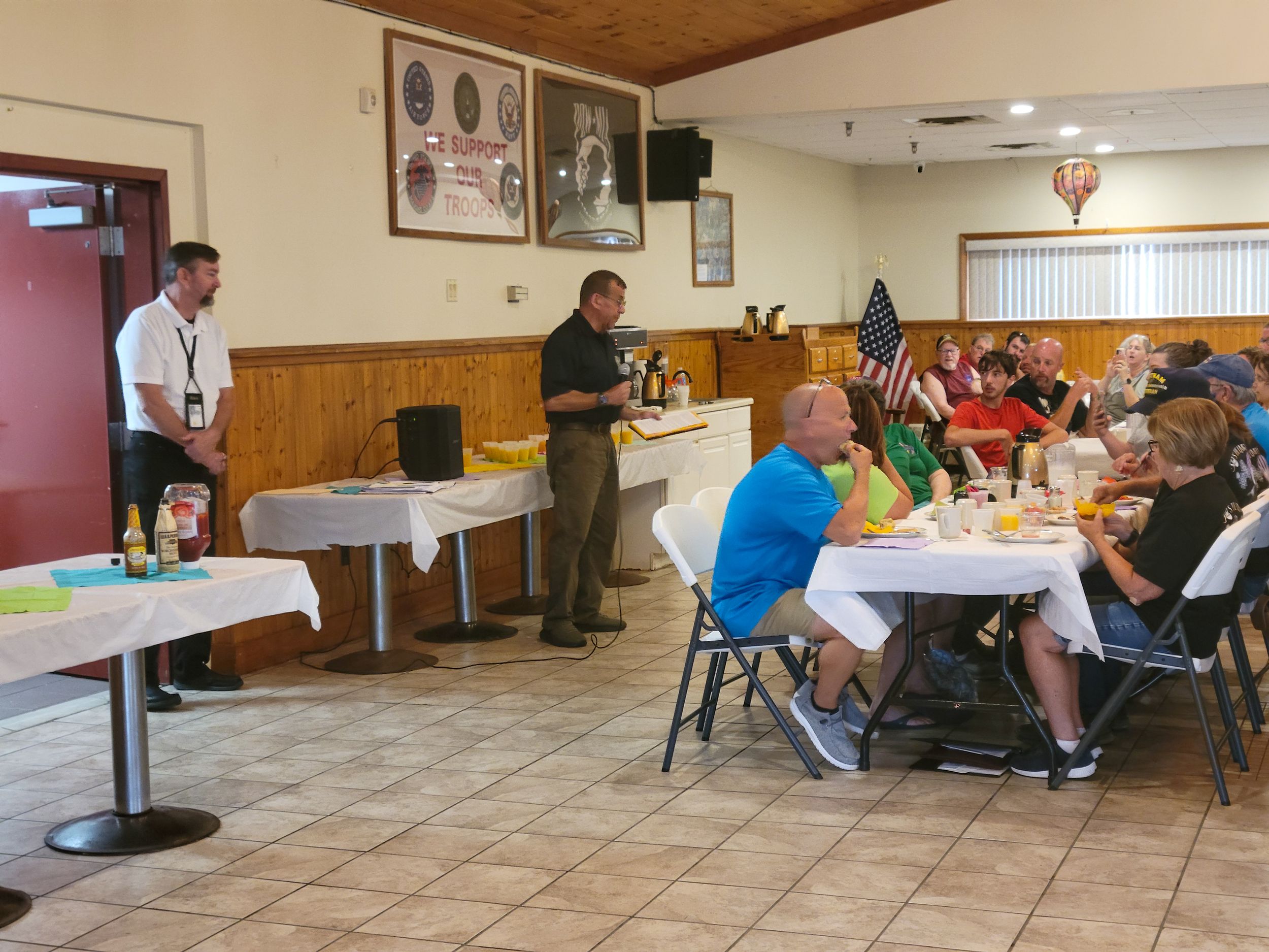 Community gathering in hall with tiled floor and wooden paneled walls.