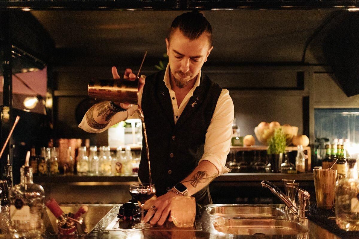 A bartender pours a cocktail while working behind a dimly lit bar counter.