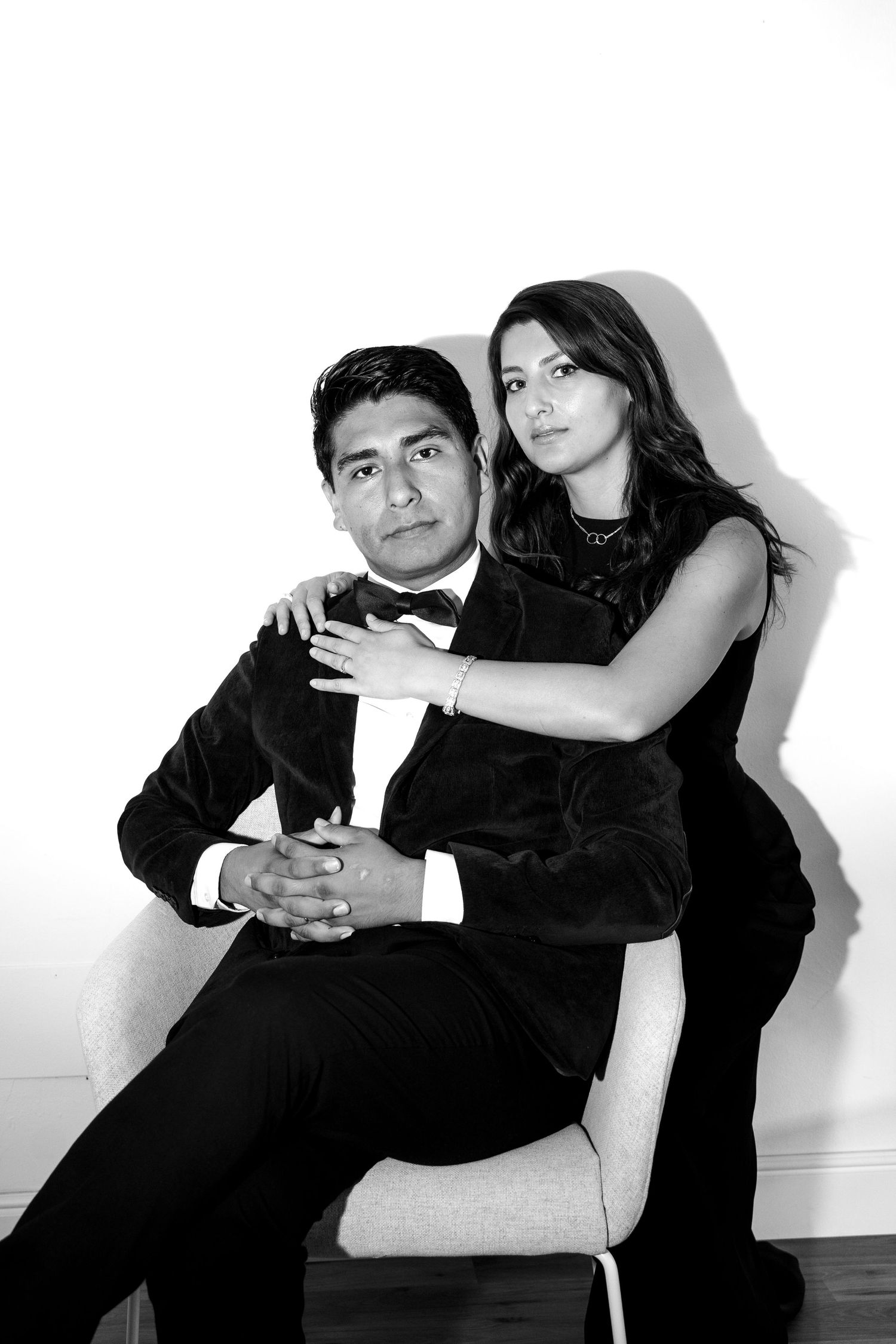 A black and white portrait of a couple in formal attire posing intimately against a white wall.