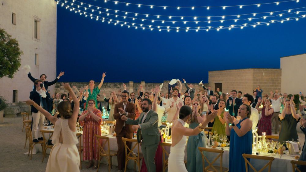 Wedding reception celebration under string lights with guests dancing and raising hands on an outdoor patio at night.