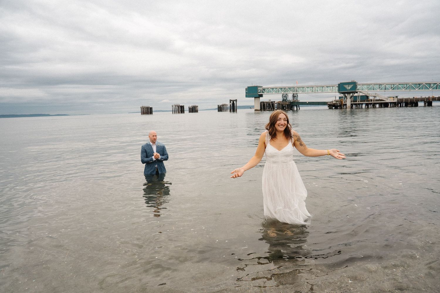 Two people in formal attire wade through ocean water near a pier on an overcast day.
