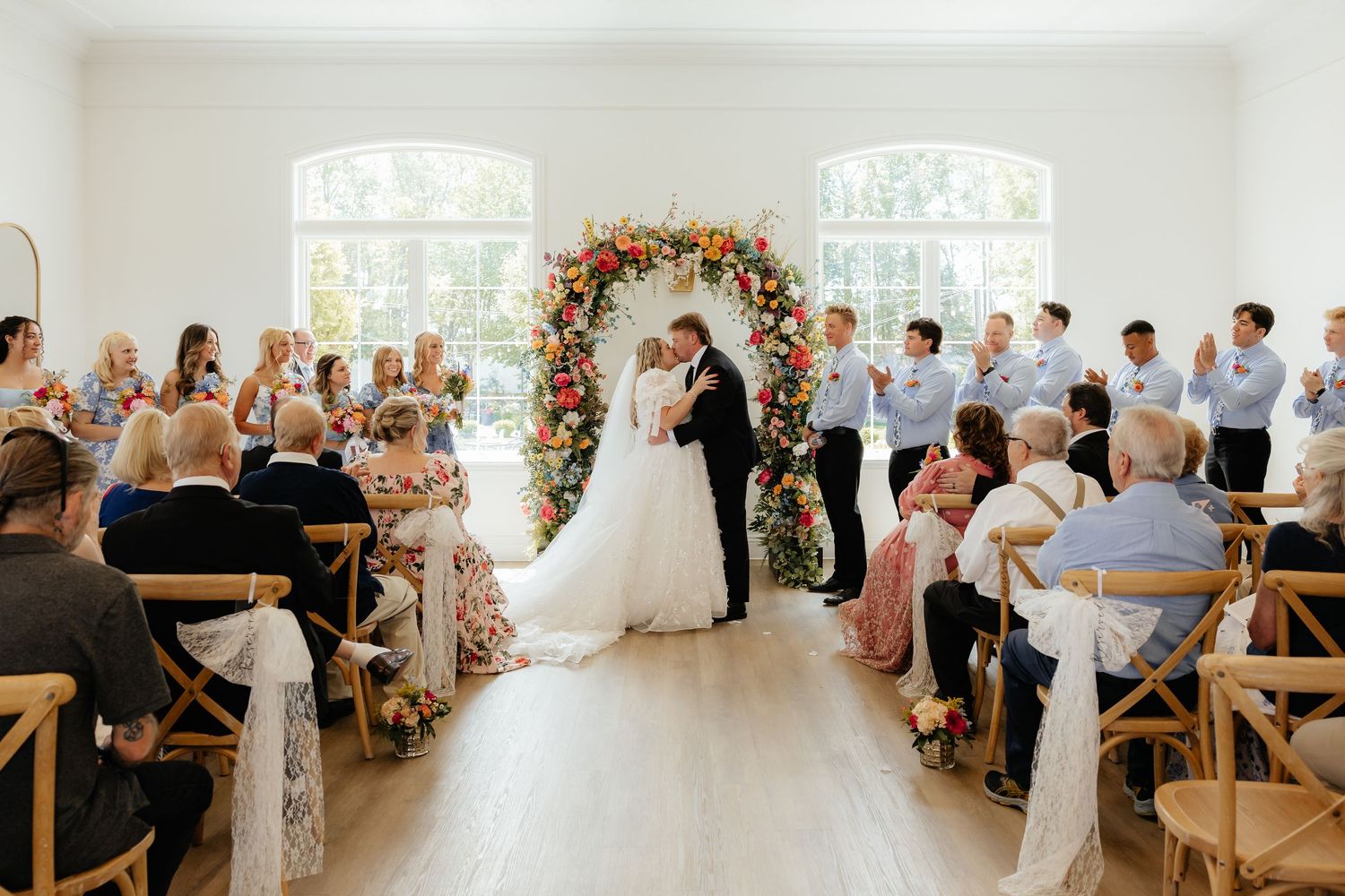 Bride and groom share first kiss at indoor wedding ceremony beneath a colorful floral arch.