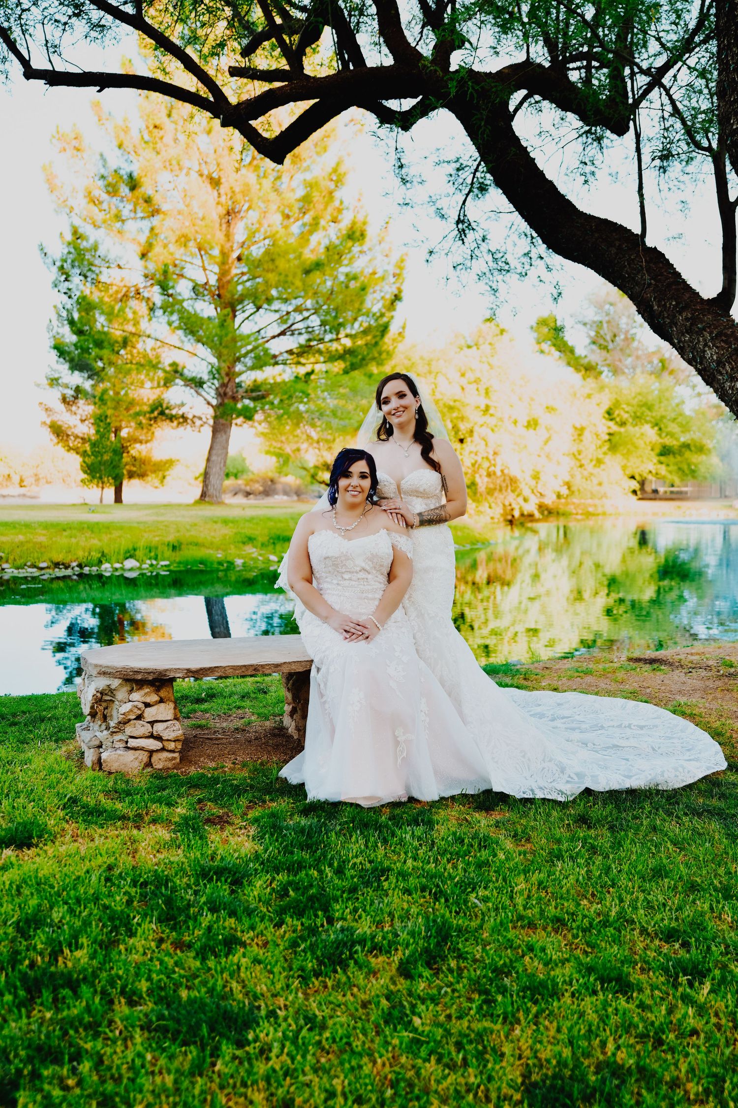 A bride in a flowing white dress sits gracefully on grass under autumn trees by a tranquil lake.