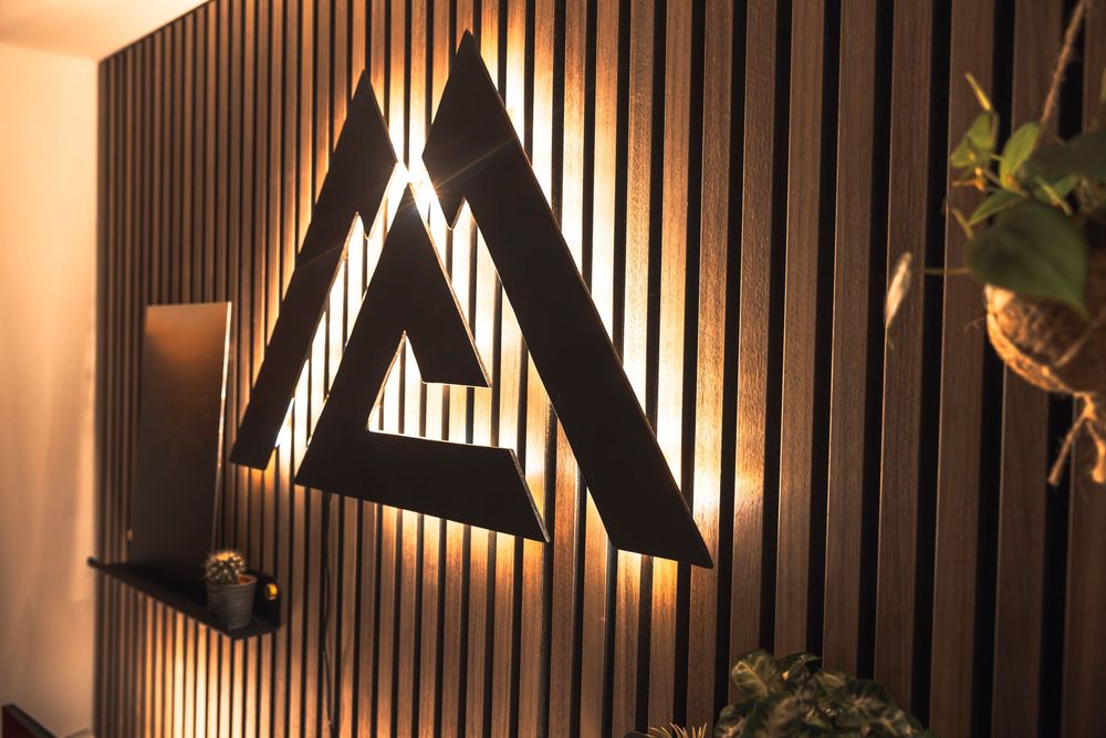 Illuminated geometric triangular logo mounted on a modern slatted wooden wall with ambient lighting and plant decor.
