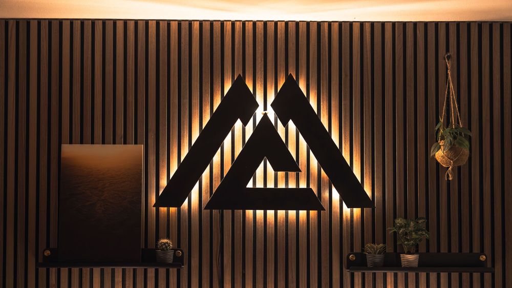 Modern illuminated triangular logo on a dark wooden slat wall with hanging plants creating an elegant ambiance.