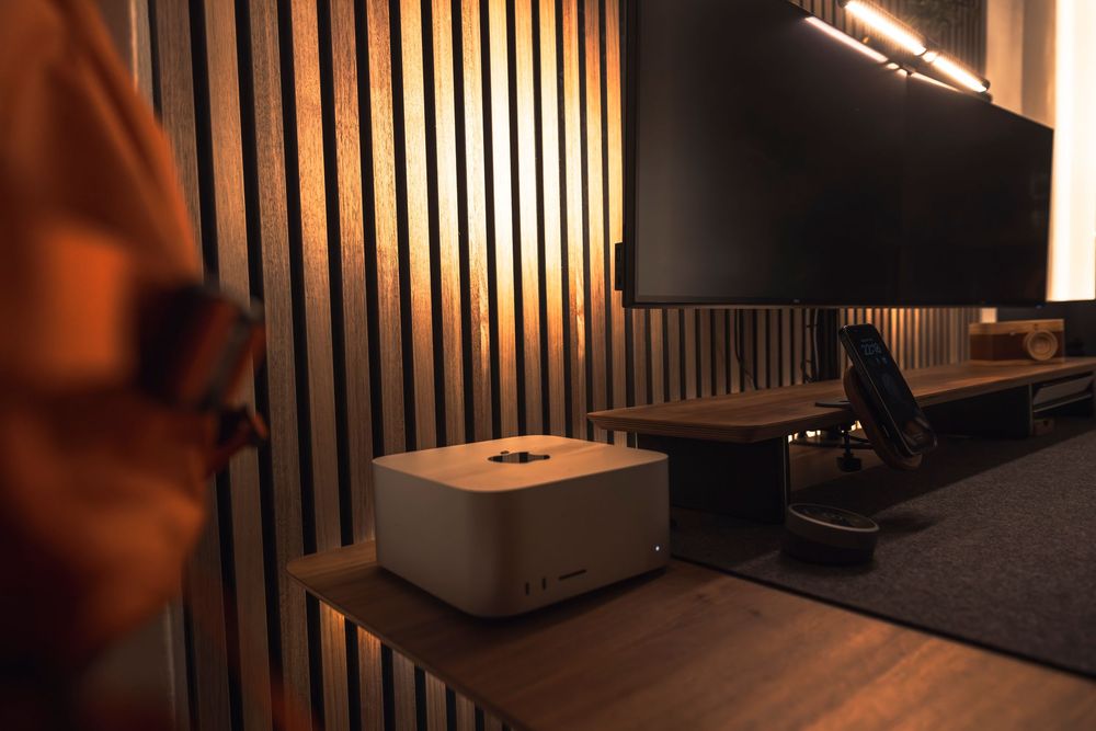 Computer equipment and tech gadgets sit on a wooden surface against a modern slatted wall with dramatic lighting.