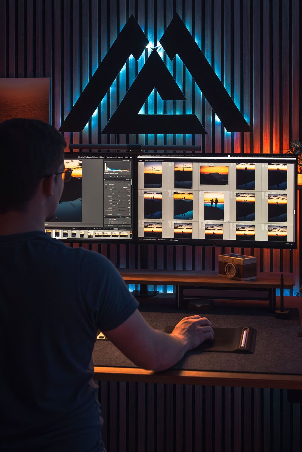 Music producer working at studio desk with illuminated triangular logo and multiple computer monitors displaying audio software.