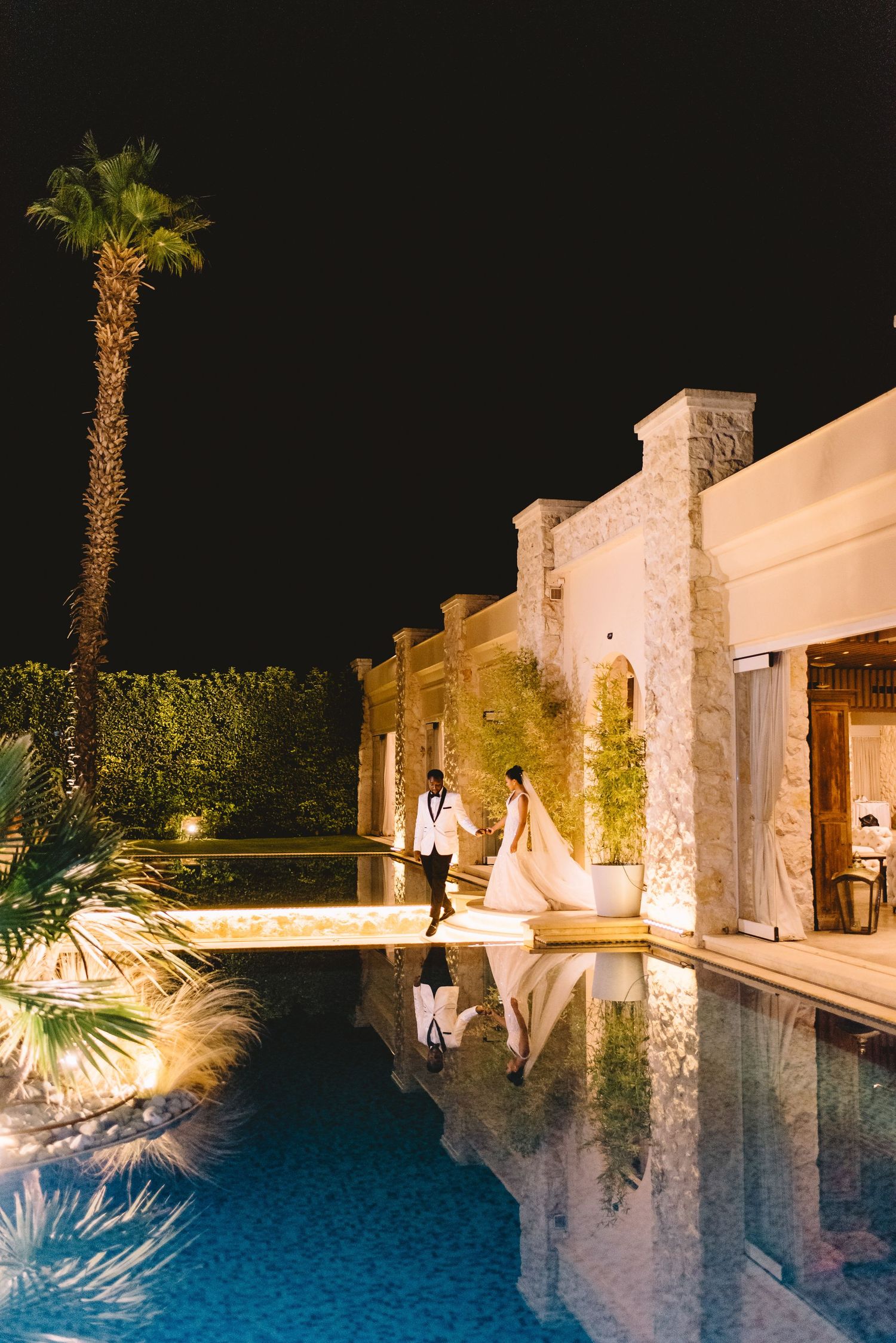 A twilight wedding scene by an illuminated swimming pool with palm trees and stone architecture creating a romantic ambiance.