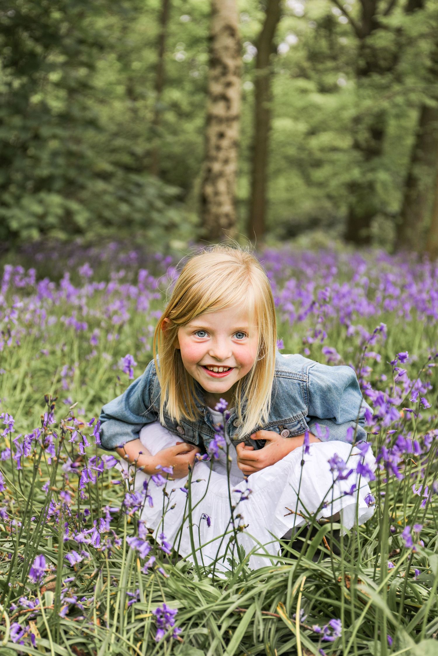 A young child kneels among vibrant purple wildflowers in a forest setting wearing denim and white clothing during family shoot in Nottingham