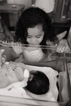 Black and white photo of a young girl with curly hair smiling at a newborn baby in a hospital bassinet.