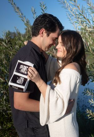 Couple sharing intimate moment while holding ultrasound photo among olive trees.
