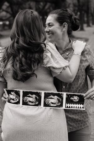 Black and white photo of two women smiling and hugging while holding ultrasound images outdoors.