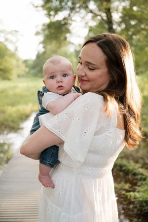 A mother in a white dress tenderly holds her baby boy outdoors on a wooden bridge surrounded by lush greenery.