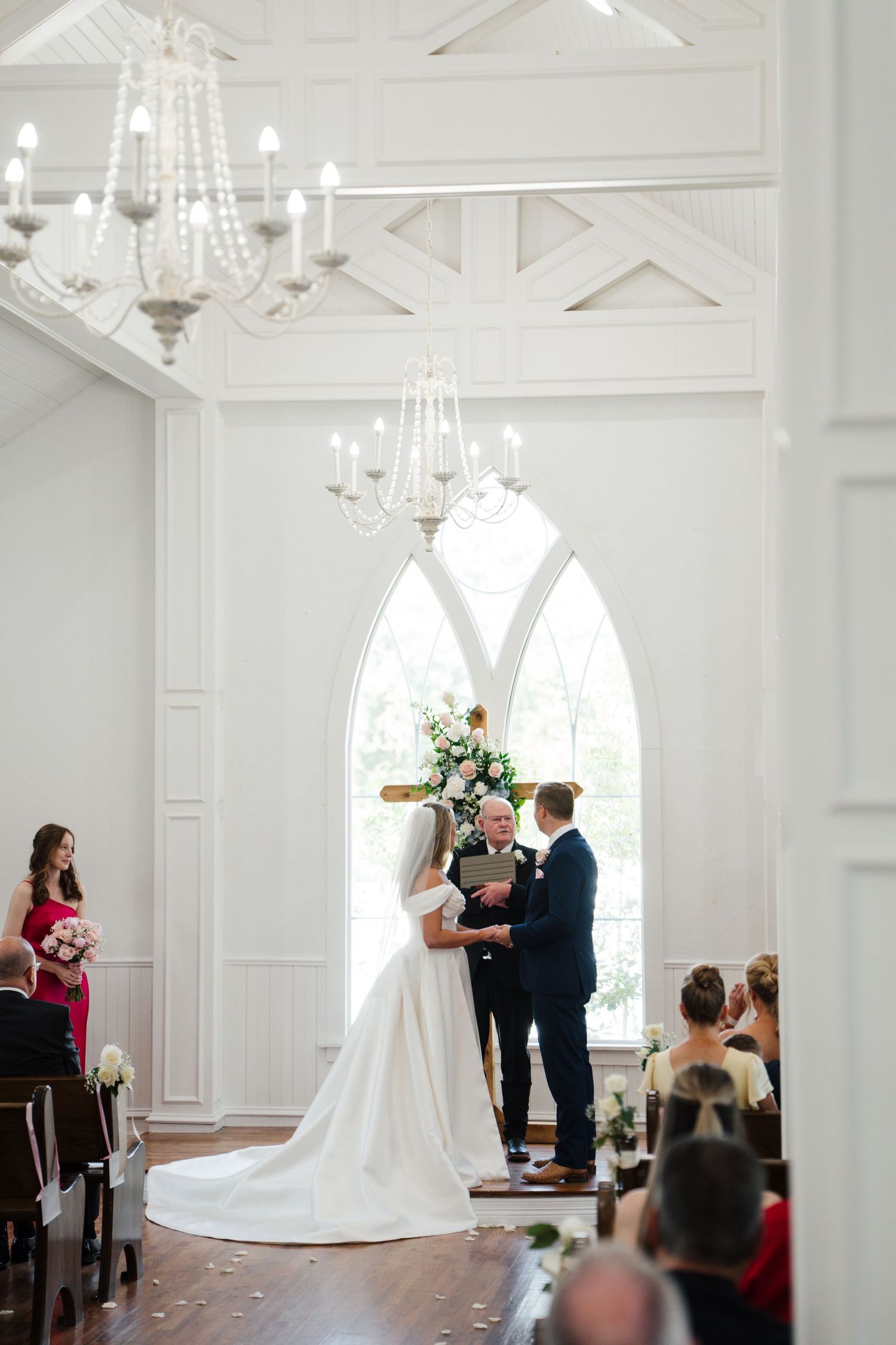 An elegant white wedding chapel interior with arched windows, crystal chandeliers and architectural details frames the ceremony.