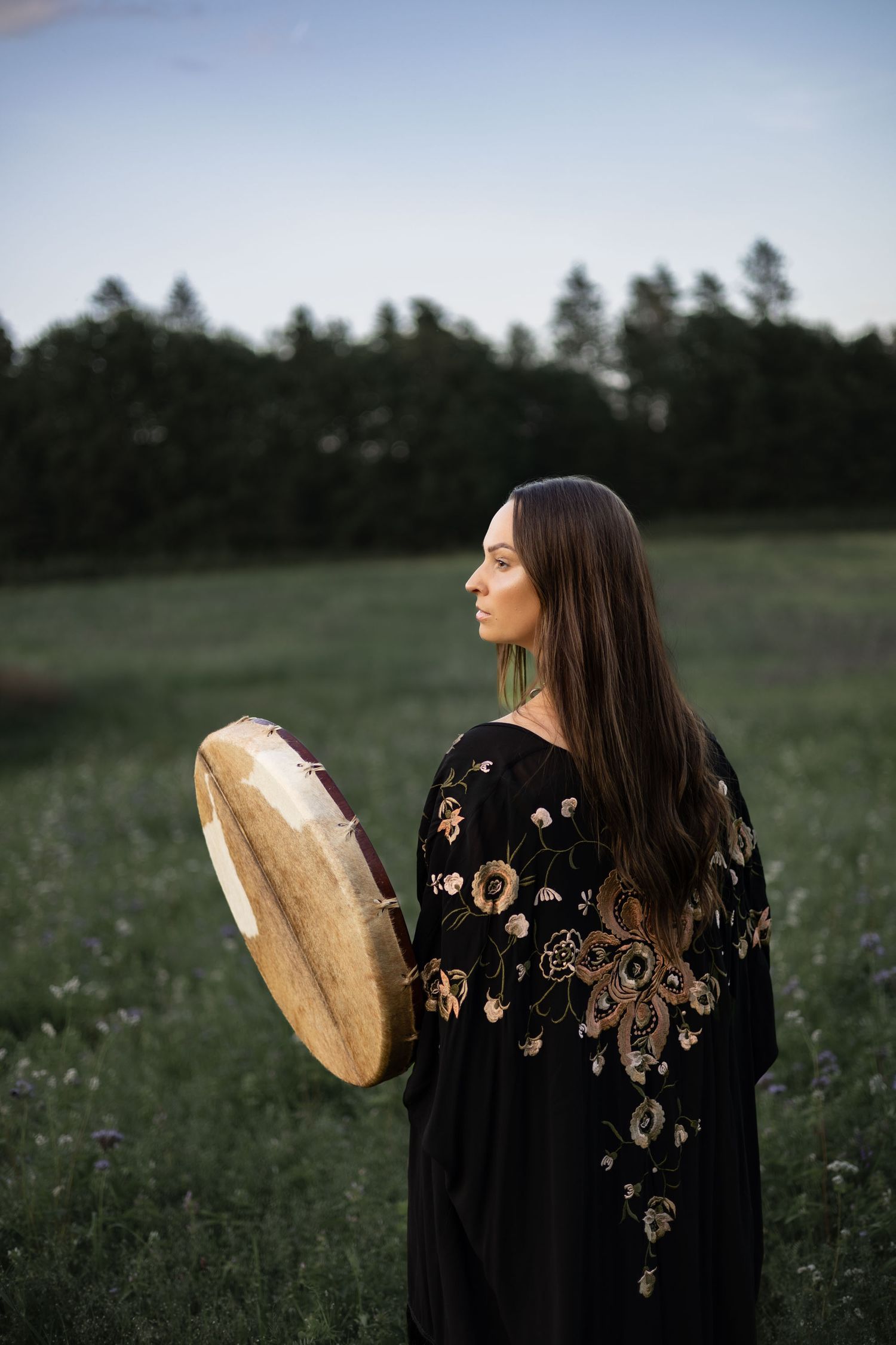 en kvinna står vänd ifrån kameran i handen har hon en trumma. på sig har hon en svart kimono med mönster. i bakgrunden syns en blå himmel.