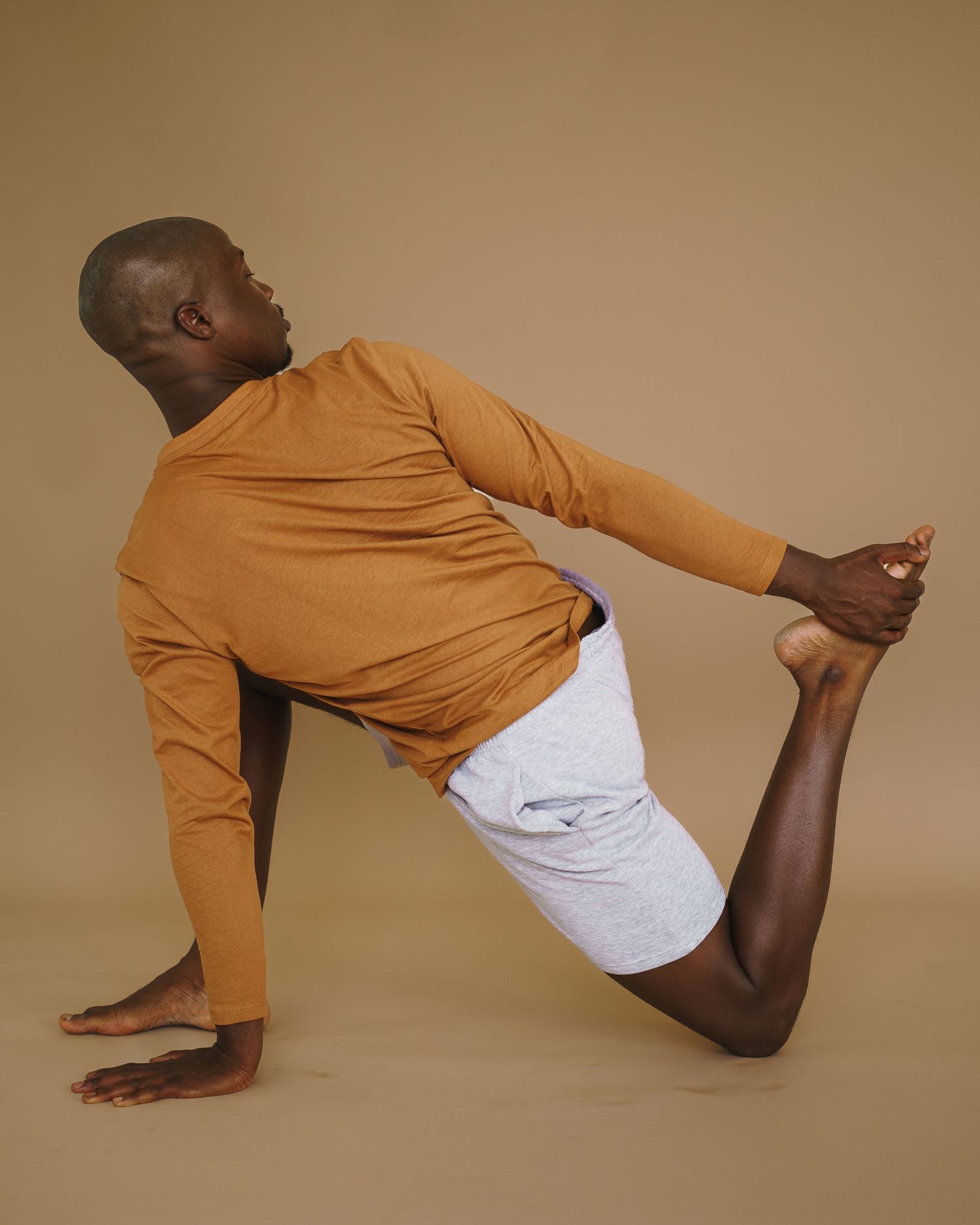 A person in an orange long sleeve top and white shorts demonstrates a dynamic yoga pose against a brown backdrop.