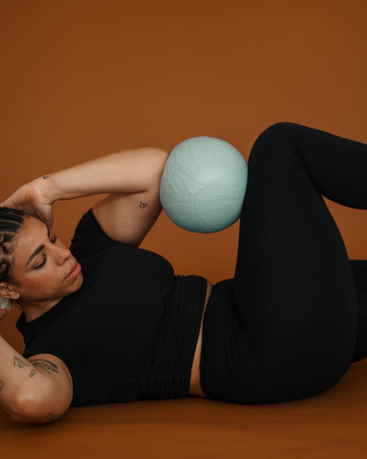 Athletic figure performs core exercise with light blue pilates ball against warm brown backdrop.