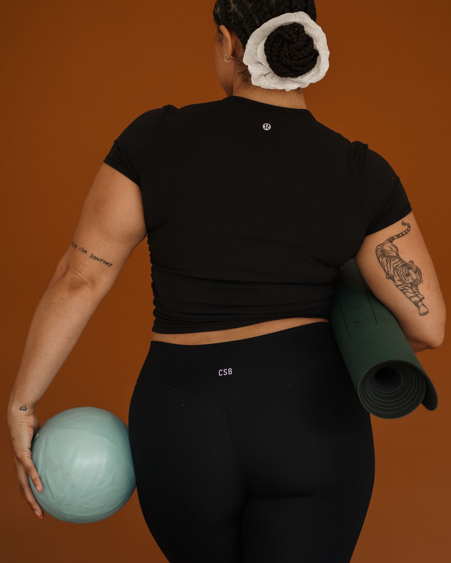 Back view of person in black workout attire holding a blue exercise ball and green yoga mat against orange backdrop.