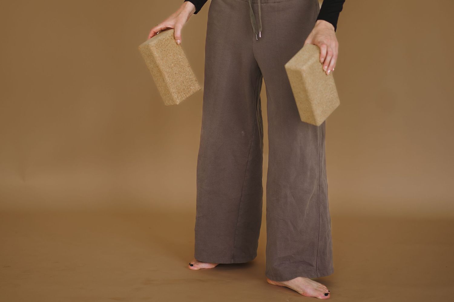 A person wearing brown wide-leg pants and black top stands with hands positioned at different angles against brown backdrop.
