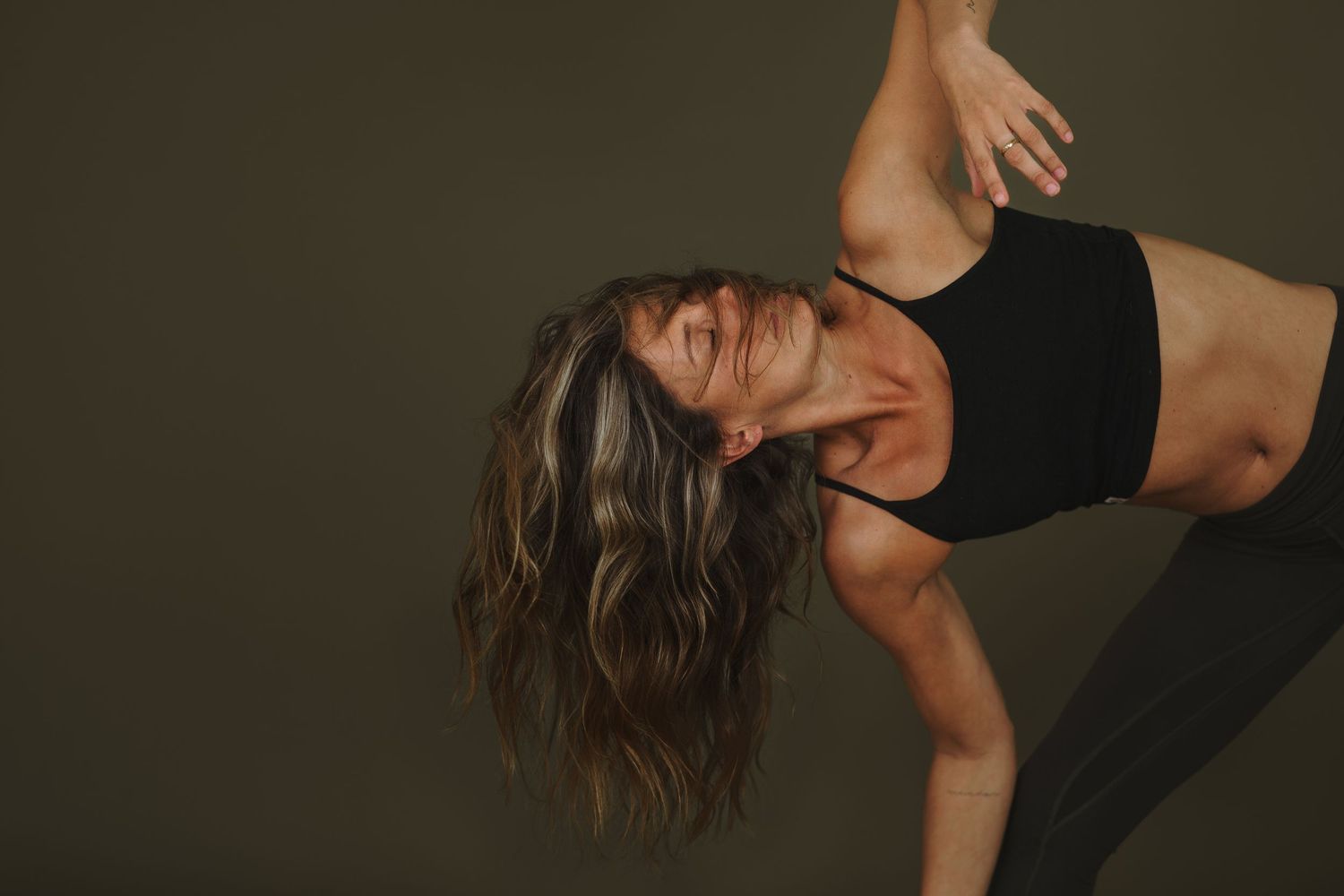 Ballet dancer in black leotard strikes graceful pose with dramatic head tilt and flowing hair against dark backdrop.