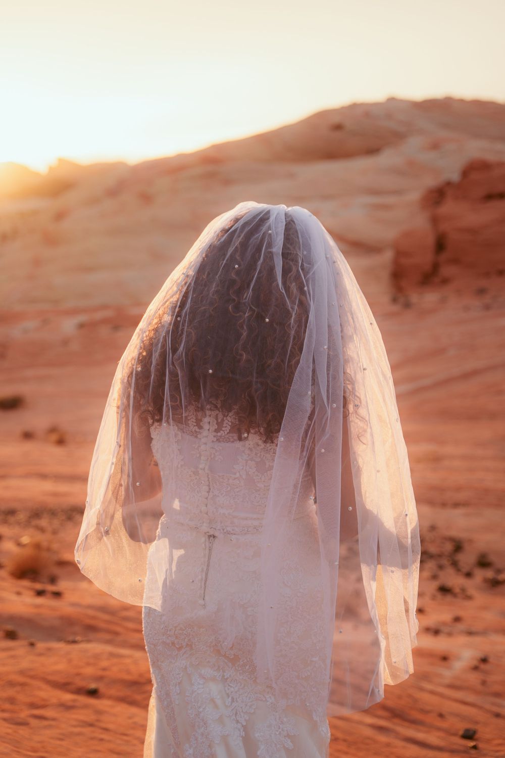 A wedding veil flows in the wind against a backdrop of red desert rock formations at sunset.