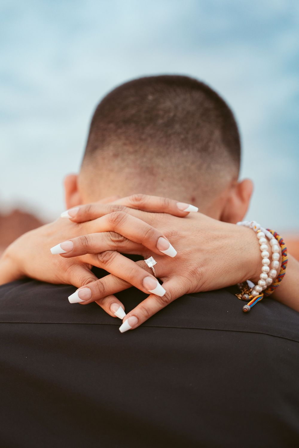 Close-up shot of hands with wedding rings resting on shoulders against a blue sky background.