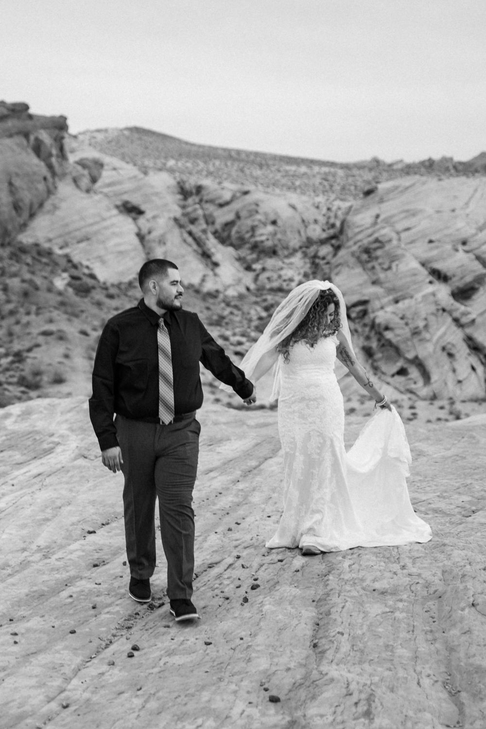 A couple in wedding attire walks hand in hand along a rocky desert landscape in black and white.