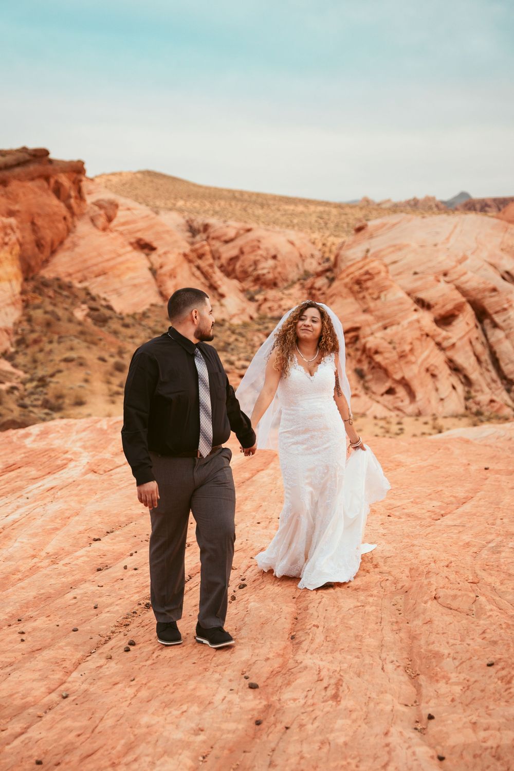 Newlyweds stroll together on red rock formations against a blue desert sky.