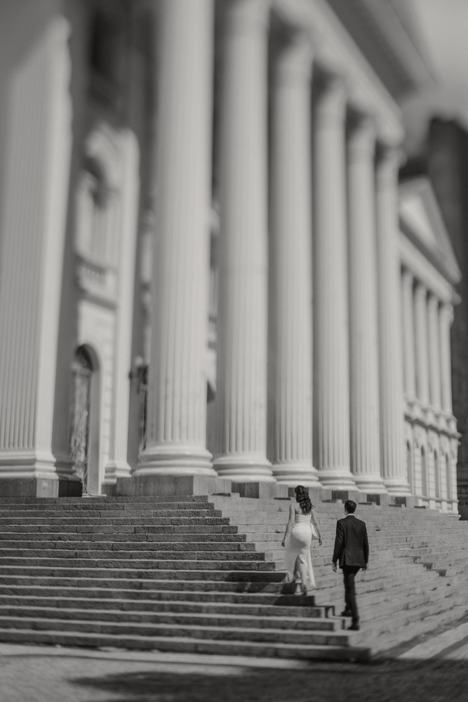 A couple ascending grand stone steps with towering classical columns in black and white photography.
