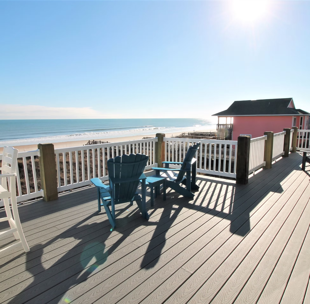 Adirondack chairs on a wooden deck overlooking a sunny beach with ocean views and a beach house with red roof.