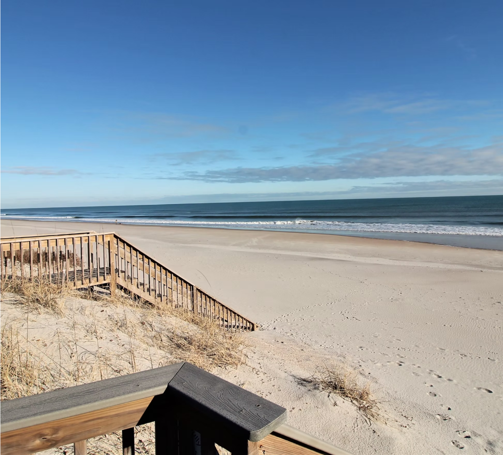 A wooden boardwalk leads down to a pristine sandy beach with blue ocean waters and clear skies on the horizon.