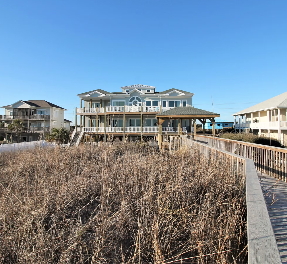 Elevated beach house with wooden boardwalk through coastal dune grass along the shoreline.