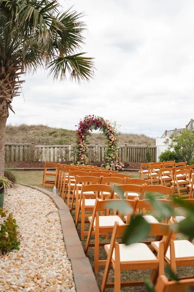A beachside wedding ceremony setup with wooden chairs and floral arch decorations along a sandy pathway flanked by palm trees.