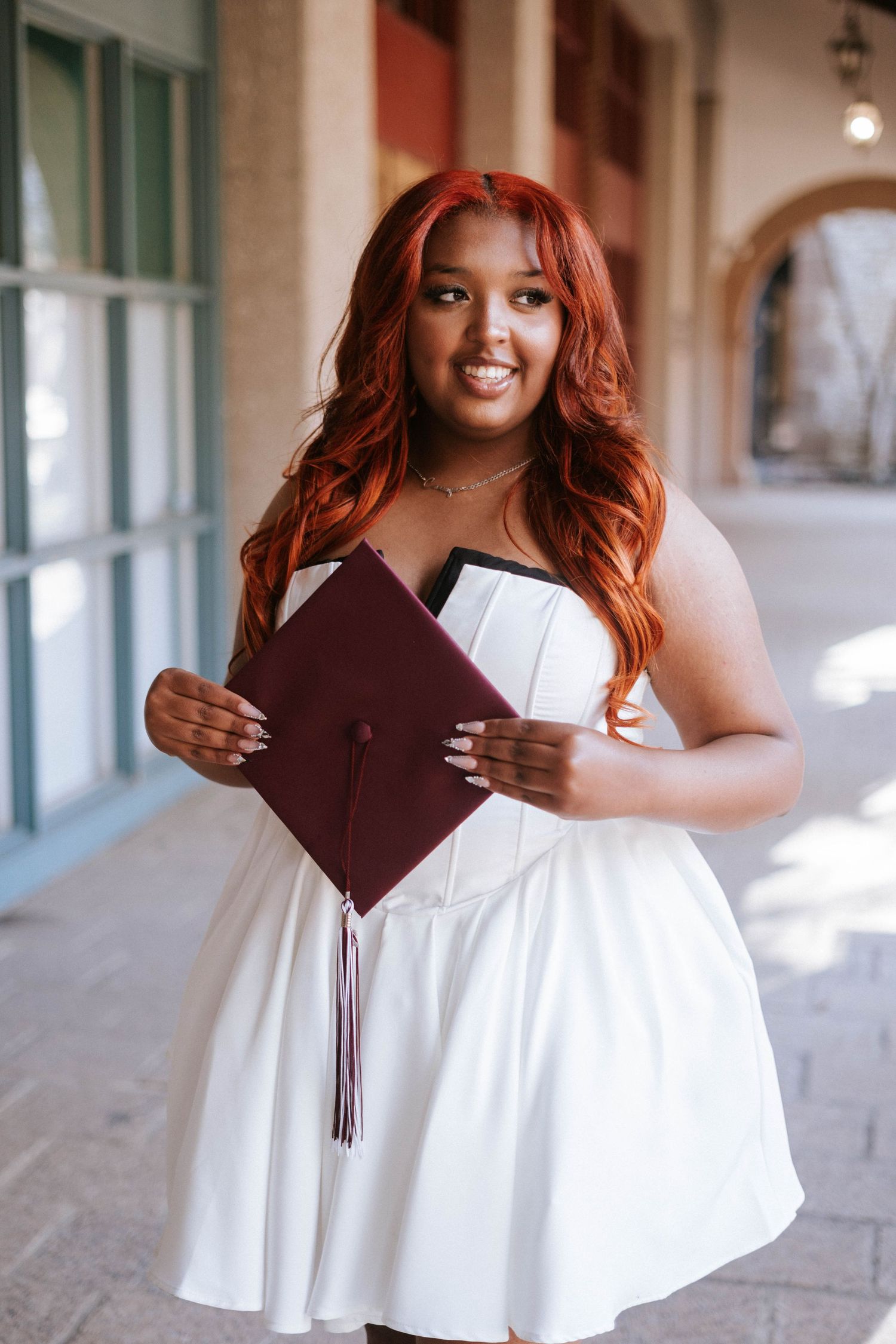 Smiling graduate in white dress with red hair holding a maroon graduation cap outdoors.