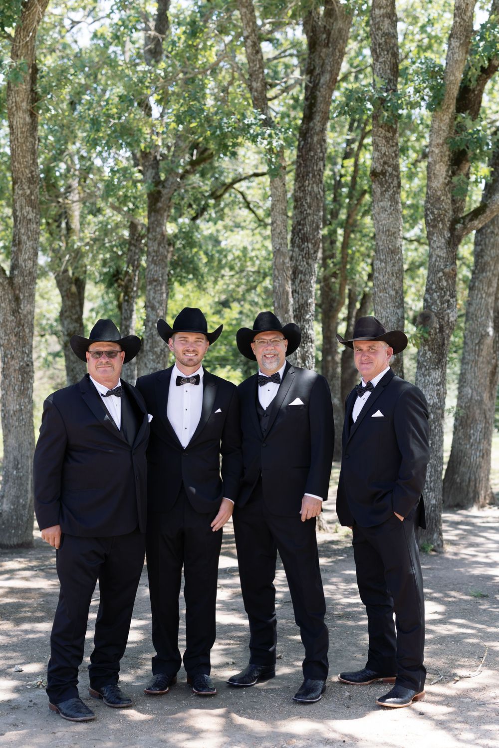 Four groomsmen in black tuxedos and cowboy hats pose together in a sunlit wooded area.