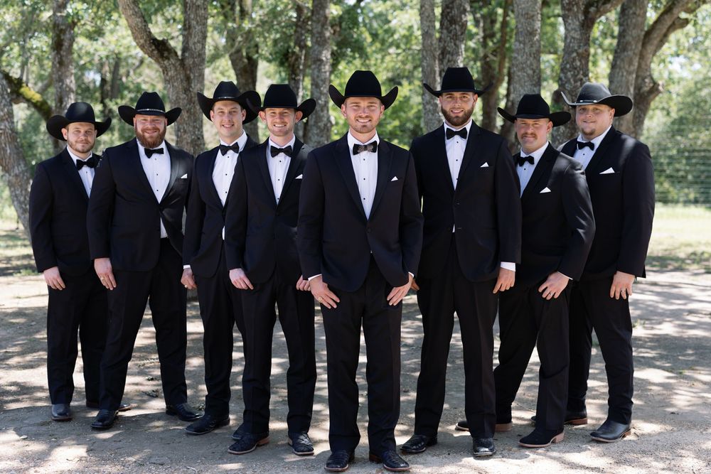 Wedding party in cowboy-themed formal wear pose together outdoors among trees.