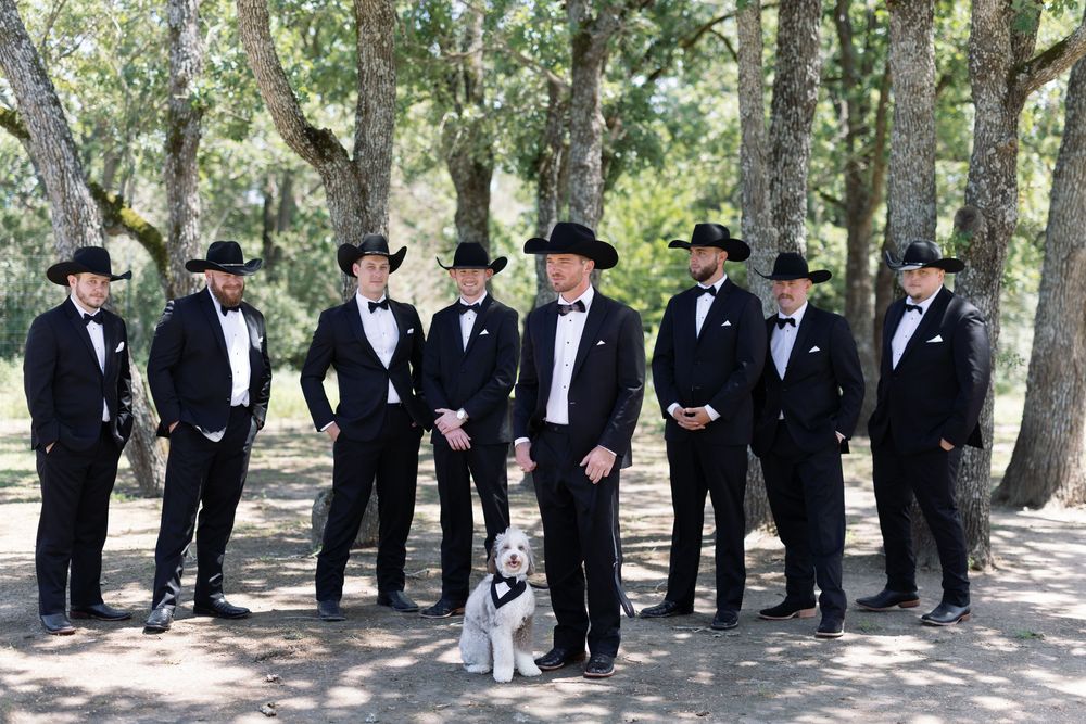 Wedding party in matching black tuxedos and cowboy hats posing with a small dog in a wooded setting.